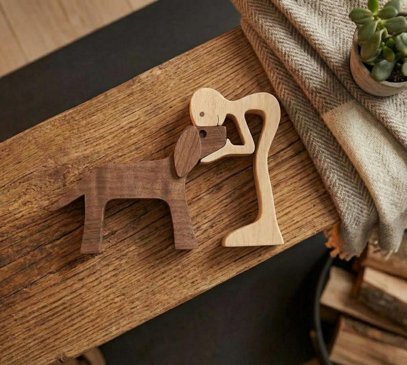 May include: Wooden dog and figure sculptures. The dog is brown, and the figure is light-colored. The figure is embracing the dog. The sculptures are on a wooden surface. A blanket and a small potted plant are in the background.