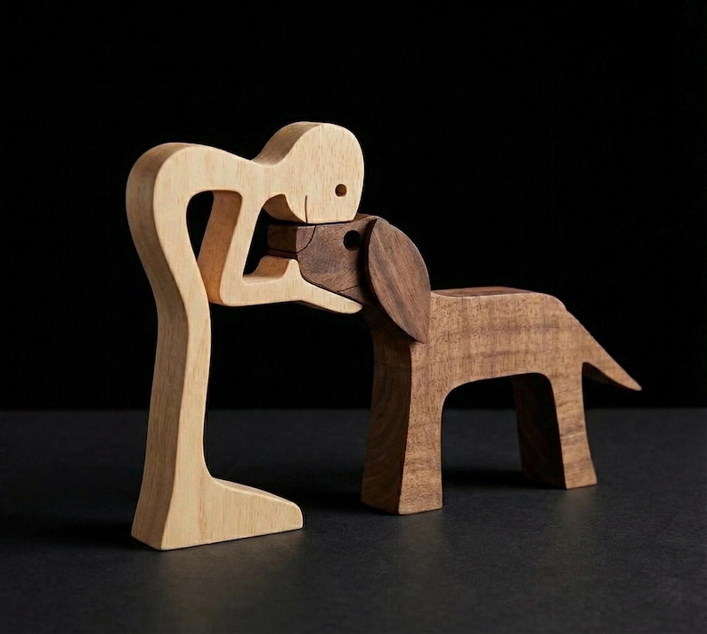 May include: A wooden sculpture of a person embracing a dog. The person is light-colored wood, while the dog is a darker wood. The sculpture is on a dark surface with a black background. The dog has large ears and a tail.
