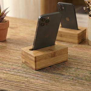 May include: Wooden phone stands with a slot to hold a smartphone. The stands are made of light-colored wood and are rectangular. Two stands are visible, each holding a black smartphone. The background includes small potted plants and a mug.