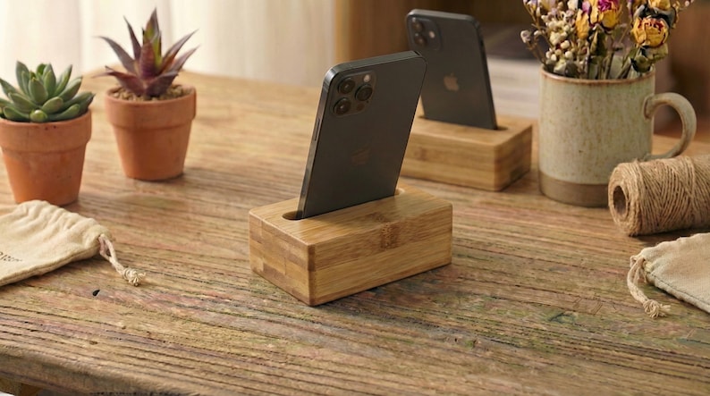 May include: Wooden phone stands with a slot to hold a smartphone. The stands are made of light-colored wood and are rectangular. Two stands are visible, each holding a black smartphone. The background includes small potted plants and a mug.