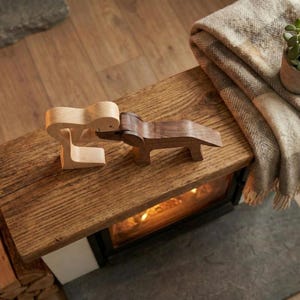 May include: Two wooden sculptures, one light-colored and one dark, are positioned on a rustic wooden surface. A folded blanket and a small potted plant are visible in the background. The sculptures appear to be abstract figures.
