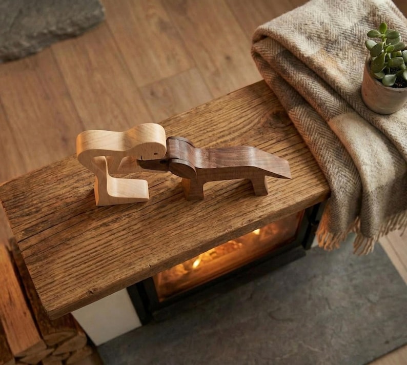 May include: Two wooden sculptures, one light-colored and one dark, are positioned on a rustic wooden surface. A folded blanket and a small potted plant are visible in the background. The sculptures appear to be abstract figures.