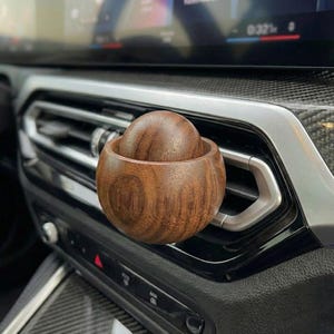 May include: A wooden car air vent diffuser with a spherical design. The diffuser is made of dark brown wood and has a matching wooden sphere inside. The diffuser is placed on a car's air vent.