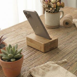 May include: A wooden phone stand holds a silver phone, angled for viewing. The stand is rectangular and made of light-colored wood. Small potted plants and a mug with dried flowers sit nearby on a rustic wooden table. A roll of twine and a small cloth bag are also present.