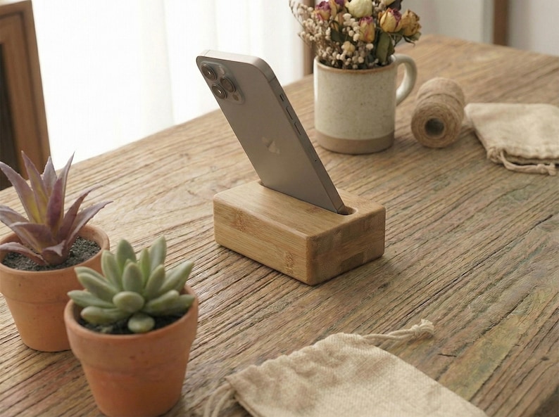 May include: A wooden phone stand holds a silver phone, angled for viewing. The stand is rectangular and made of light-colored wood. Small potted plants and a mug with dried flowers sit nearby on a rustic wooden table. A roll of twine and a small cloth bag are also present.