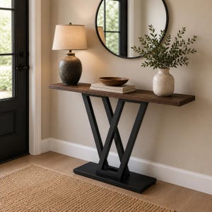 Può includere: Un tavolo console con piano in legno marrone scuro e base in metallo nero con struttura di supporto a V. Il tavolo sostiene una lampada scura testurizzata con paralume beige, una ciotola in legno su libri impilati e un vaso bianco con fogliame.