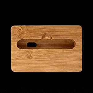 May include: A rectangular bamboo phone stand with a slot for a phone and a small hole for a charging cable. The stand has a natural wood grain finish and a small handle on the top. The stand is on a black background.