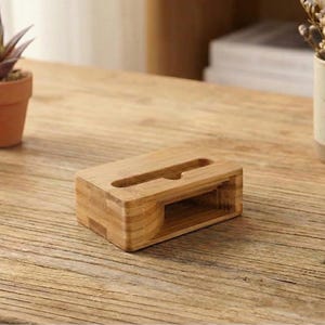 May include: A wooden phone stand with a slot for a phone and a lower shelf. The stand is made of light-colored wood and sits on a wooden table. Small potted plants and a mug with dried flowers are also on the table.