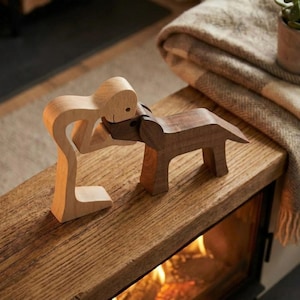May include: Wooden figurine of a person embracing a dog. The person is light-colored wood, and the dog is a darker wood. The figures are on a wooden surface, with a blanket and plant in the background. The scene evokes warmth and affection.