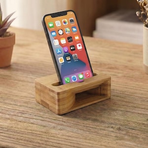 May include: A wooden phone stand holds a smartphone with a colorful app display. The stand is made of light-colored wood and has a rectangular shape with a slot for the phone. Small potted plants and a mug with dried flowers are in the background.