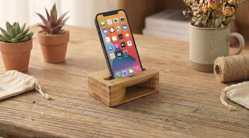 May include: A wooden phone stand holds a smartphone with a colorful app display. The stand is made of light-colored wood and has a rectangular shape with a slot for the phone. Small potted plants and a mug with dried flowers are in the background.