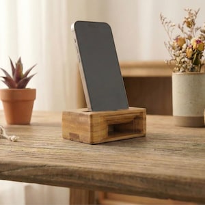 May include: A wooden phone stand with a slot to hold a smartphone. The stand is made of light-colored wood and sits on a rustic wooden table. The phone is black with a silver frame. Decorative plants and a mug are in the background.
