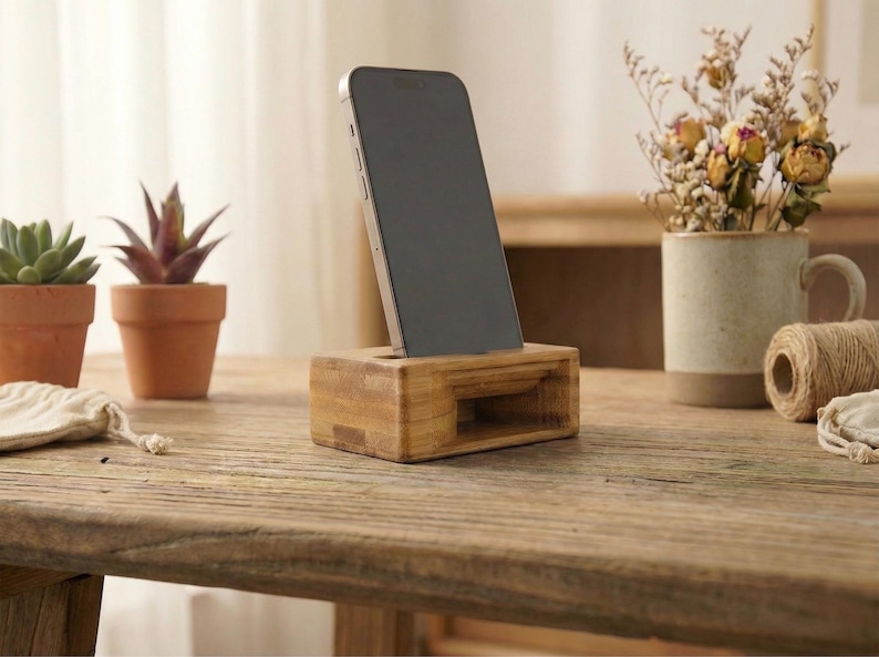 May include: A wooden phone stand with a slot to hold a smartphone. The stand is made of light-colored wood and sits on a rustic wooden table. The phone is black with a silver frame. Decorative plants and a mug are in the background.
