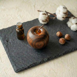 以下が含まれることがあります： 木製のリンゴが、小さな琥珀色のガラス瓶の隣の濃い灰色のスレートの上に置かれています。3つのヘーゼルナッツと綿の小枝もスレートにあります。リンゴは天然の木目を持っています。