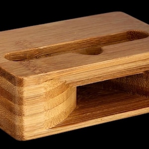 May include: A rectangular bamboo phone stand with a slot for a phone and a smaller hole. The stand is made of layered bamboo and has a natural wood grain finish. The stand is designed to hold a phone upright.