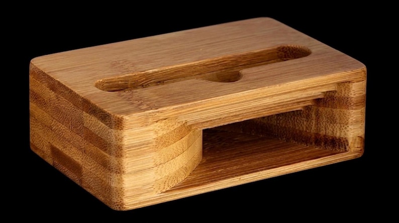 May include: A rectangular bamboo phone stand with a slot for a phone and a smaller hole. The stand is made of layered bamboo and has a natural wood grain finish. The stand is designed to hold a phone upright.