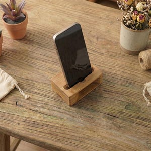 May include: A wooden phone stand holds a black smartphone on a rustic wooden table. Small potted plants and a mug with dried flowers are also on the table. A small roll of twine and two small drawstring bags are also present.