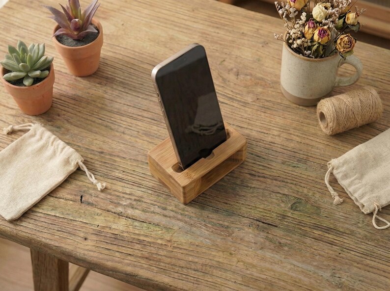 May include: A wooden phone stand holds a black smartphone on a rustic wooden table. Small potted plants and a mug with dried flowers are also on the table. A small roll of twine and two small drawstring bags are also present.