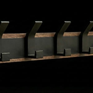 Pu&ograve; includere: Un attaccapanni rustico da parete con cinque ganci in metallo scuro. I ganci sono montati su una tavola rettangolare di legno con un centro marrone scuro e un bordo in legno pi&ugrave; chiaro. Design semplice e moderno.