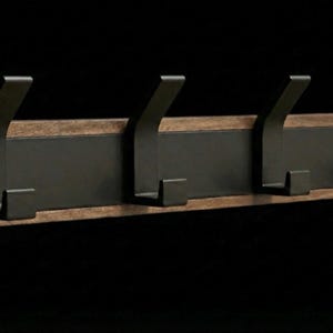 Pu&ograve; includere: Un attaccapanni da parete con quattro ganci in metallo scuro. I ganci sono montati su una tavola rettangolare di legno con un pannello centrale in metallo scuro. Il legno ha una venatura naturale e un caldo tono marrone.