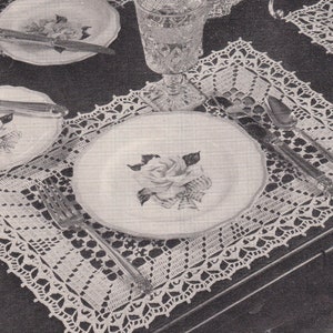 May include: A black and white photo of a table setting with four place settings. Each place setting has a white crocheted doily, a white plate with a floral design, a silver knife and fork, and a glass. There is a silver serving dish with a lid in the center of the table.
