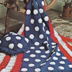 May include: A blue and white crochet blanket with white daisy-like flowers, a red and white striped crochet blanket, and a white crochet pillow with white daisy-like flowers. The blankets are on a patterned rug with a spinning wheel and a vase of branches in the background. The text "stars and stripes" is at the top of the image.