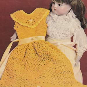May include: A crocheted doll dress in a yellow color with a white satin ribbon. The dress has a scalloped edge and a crocheted flower design.