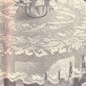 May include: A white crocheted tablecloth with a delicate floral pattern. The tablecloth is laid over a round table and has a scalloped edge.