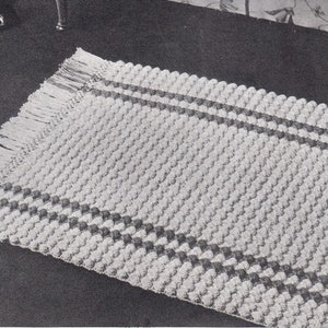 Puede incluir: Una alfombra blanca de crochet con una superficie texturizada y dos rayas grises oscuras. La alfombra tiene un borde de flecos.