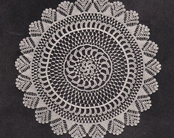 Puede incluir: Un tapete de ganchillo circular, de color blanquecino, con un intrincado diseño de encaje. El diseño presenta círculos concéntricos y un borde festoneado con detalles en forma de hojas. Sobre un fondo oscuro.
