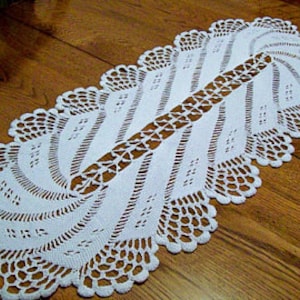May include: White crocheted table runner with a delicate, intricate pattern. The runner is oval shaped and has a scalloped edge.