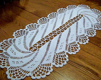 Puede incluir: Un camino de mesa de crochet blanco con un patrón delicado e intrincado. El camino de mesa es de forma ovalada y tiene un borde festoneado.