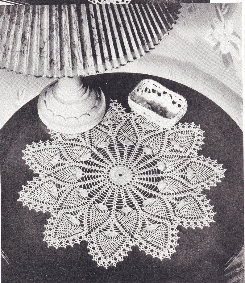 May include: A white crocheted doily with a peacock feather design. The doily is on a dark surface with a lamp and a small decorative box.