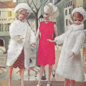 May include: Three dolls wearing knitted clothing. The doll on the left is wearing a white coat and a brown skirt. The doll in the middle is wearing a red dress. The doll on the right is wearing a white coat and a brown skirt.