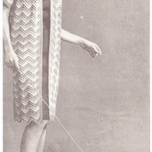 May include: A woman wearing a white crocheted cardigan with a chevron pattern and a black dress is walking a small white dog wearing a crocheted sweater. The dog is on a leash.