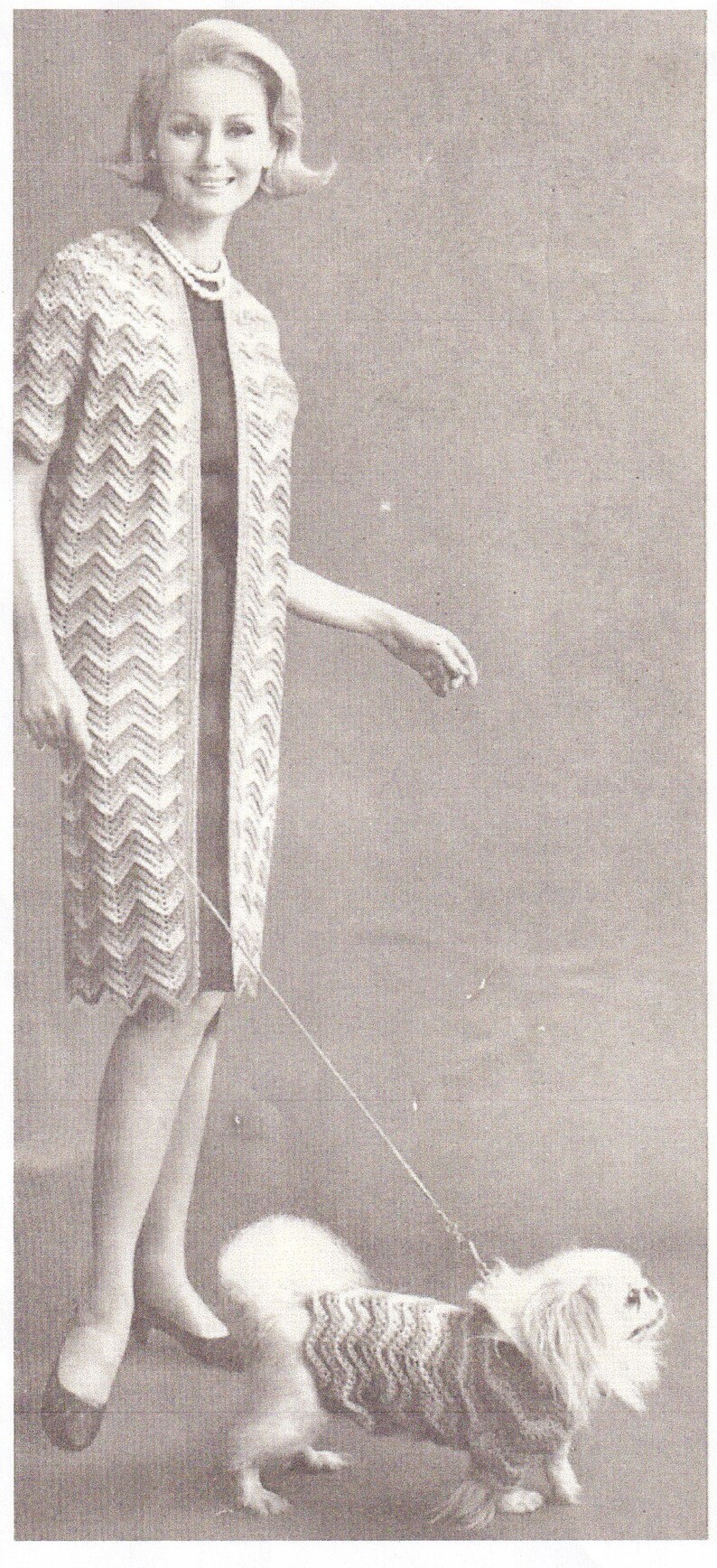 May include: A woman wearing a white crocheted cardigan with a chevron pattern and a black dress is walking a small white dog wearing a crocheted sweater. The dog is on a leash.