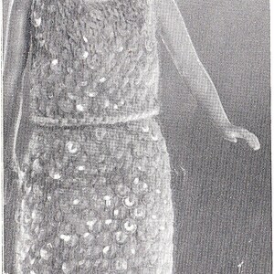 May include: A black and white photo of a doll wearing a crocheted dress. The dress has a fitted bodice and a flowing skirt.