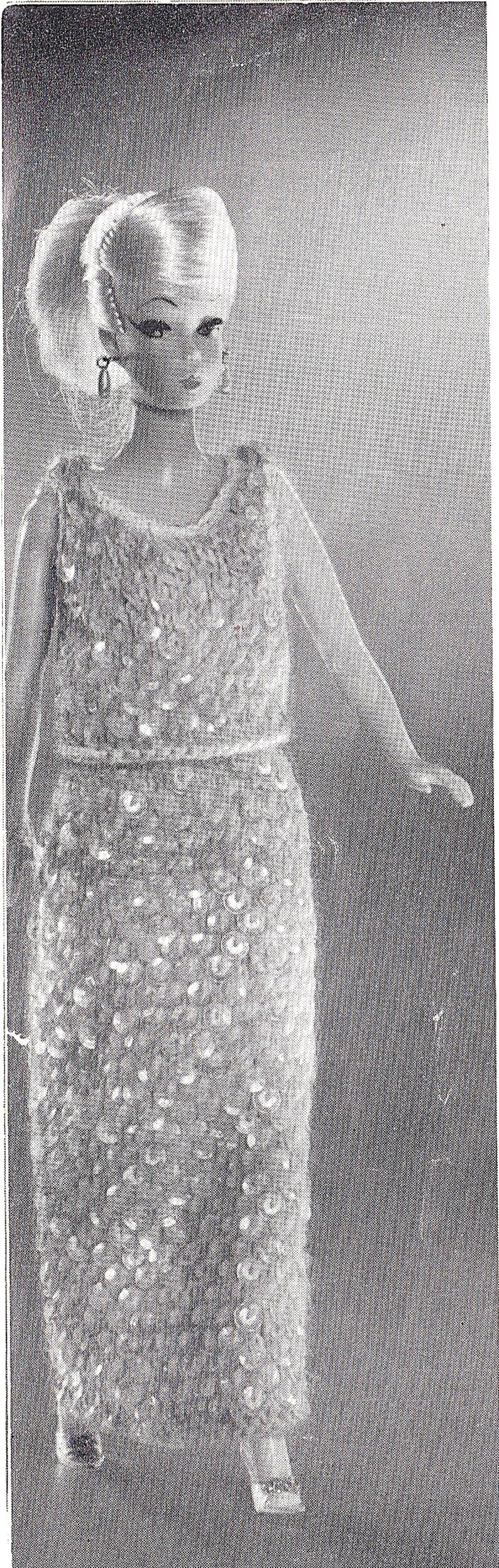 May include: A black and white photo of a doll wearing a crocheted dress. The dress has a fitted bodice and a flowing skirt.