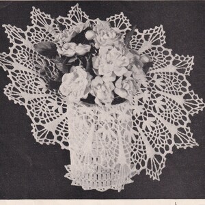 May include: A white, crocheted basket filled with white roses, set on a matching doily. The basket and doily feature intricate openwork patterns. The image is a close-up, showcasing the delicate details of the handmade craft.