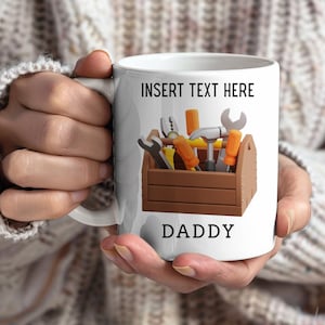Puede incluir: Taza de cerámica blanca con un gráfico de caja de herramientas marrón llena de herramientas. La taza tiene el texto "INSERT TEXT HERE" en la parte superior y "DADDY" en la parte inferior. La taza está sujeta por una persona.