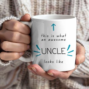 May include: White ceramic mug with the text "this is what an awesome UNCLE looks like" in black and teal lettering. An upward-pointing arrow and decorative lines accent the text. The mug is held by a person.