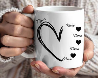 Könnte beinhalten: Weiße Keramiktasse mit einem schwarzen Herzdesign und dem Wort "Mom". Unter dem Herzen stehen die Worte "Name" und drei schwarze Herzsymbole. Die Tasse wird von einer Person gehalten.