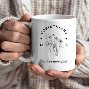 Puede incluir: Taza de cerámica blanca con el texto "CORINTHIANS 13:8" y una ilustración floral. La frase "His love never fails" está escrita debajo. La taza tiene un asa curva.