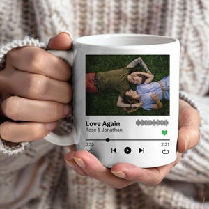 May include: White ceramic mug featuring a photo of two people lying on grass, with the text "Love Again" and a music player interface. The mug has a handle and is held by a person.