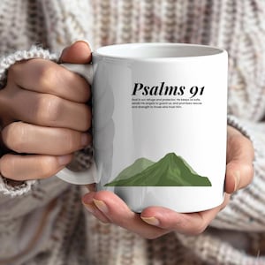 Puede incluir: Taza de cerámica blanca con el texto "Psalms 91" en negro, una cita y un gráfico de montaña verde. La taza está sostenida por una persona.
