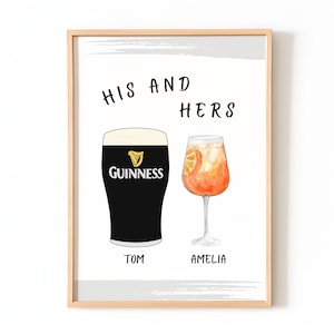 Op de afbeelding: Een ingelijste print met de tekst "HIS AND HERS" boven illustraties van een pint Guinness en een cocktail. Het Guinness-glas is zwart met het Guinness-logo. De cocktail is oranje met een sinaasappelschijf. De namen "TOM" en "AMELIA" staan eronder.