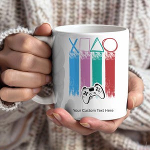 Op de afbeelding: Witte keramische mok met een gaming-thema. Het heeft een gamecontroller en de tekst "Your Custom Text Here". Het ontwerp bevat blauwe, paarse, turquoise en rode verticale penseelstreken met gamecontroller-symbolen.