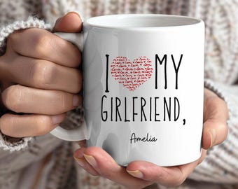 Op de afbeelding: Witte keramische mok met de tekst "I ❤️ MY GIRLFRIEND," en de naam "Amelia." Het hart is gevuld met het woord "love" in rood. De mok wordt door een persoon vastgehouden.