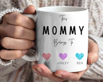 Könnte beinhalten: Weiße Keramik-Tasse mit dem Text "This MOMMY Belongs To" in schwarzer Schrift. Darunter drei Herzen in Rosa, Lila und Blau, mit den Namen Lily, Ashley und Ben. Die Tasse hat einen Henkel.