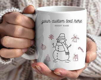 May include: White ceramic mug featuring a snowman illustration with a hat and scarf, surrounded by festive doodles. The mug has the text "your custom text here" and "INSERT NAME" in black script.